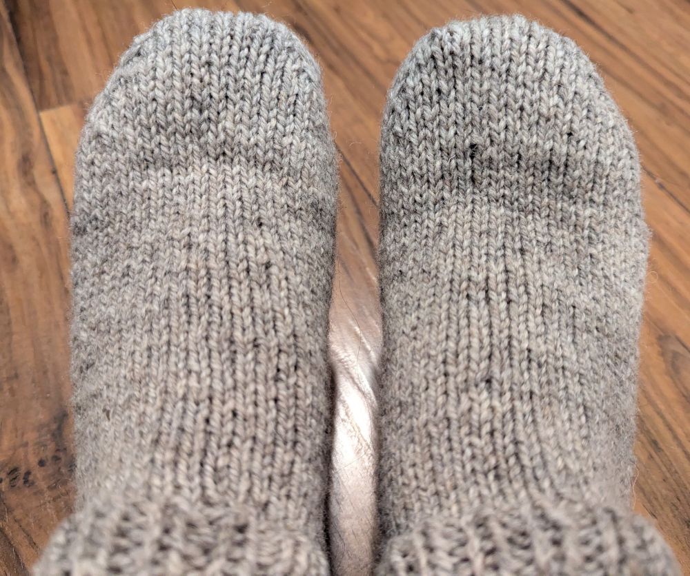 A pair of feet clad in two oatmeal-coloured short socks known as 'vamps.' The feet are resting on a brown hardwood floor.