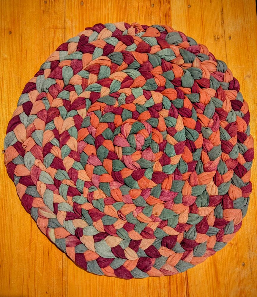 A grey, burgundy, and red braided rug, sewn into a circle shape. It lays on a wood table.
