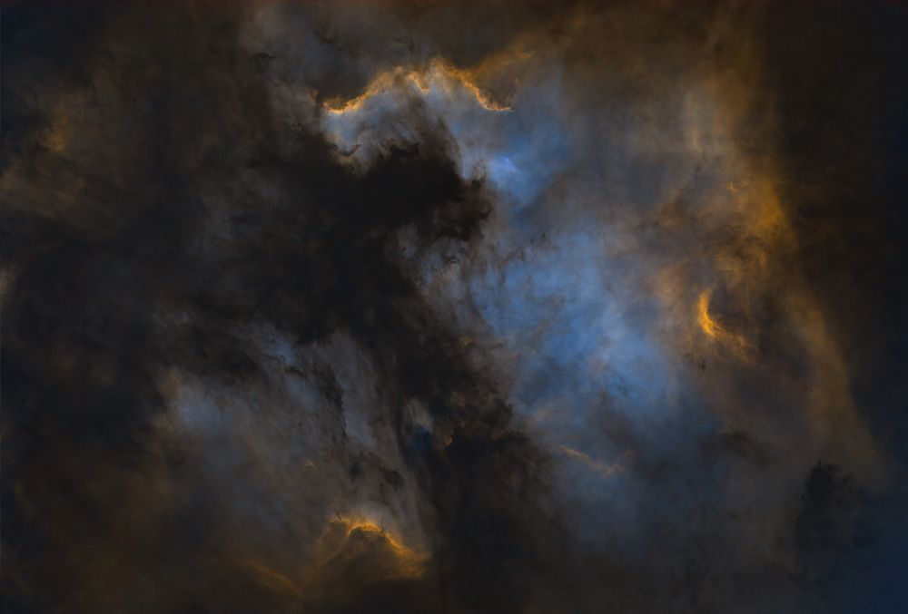 A swirl of gasses in space, represented with sapphire and gold colors. A black void between two nebulae resembles a cross whose limbs are lobed. Stars removed, the image resembles an oil painting.