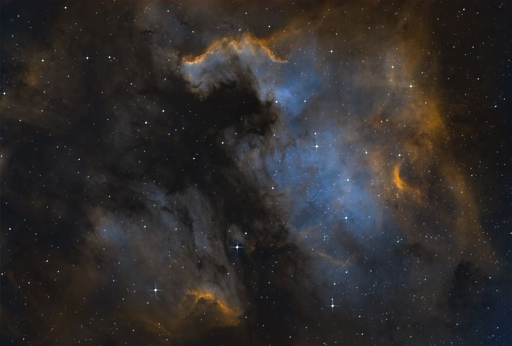 A swirl of gasses in space, represented with sapphire and gold colors. A black void between two nebulae resembles a cross whose limbs are lobed. The image is peppered with stars.