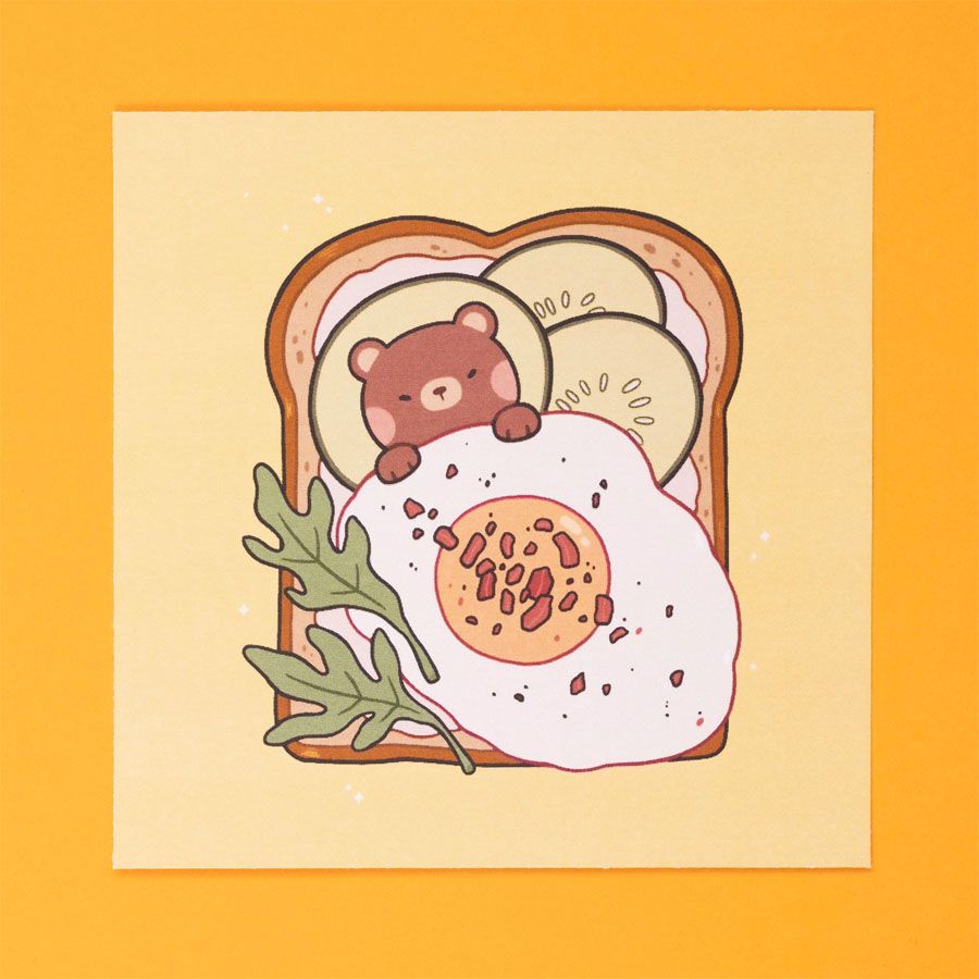 Art print titled "Bear and Breakfast", depicting a small brown bear sleeping atop a slice of buttered toast, with cucumber slices for pillows and a sunny-side-up for a blanket. A couple of rocket leaves can be seen on the bottom side, and there are bits of bacon over the egg blanket.