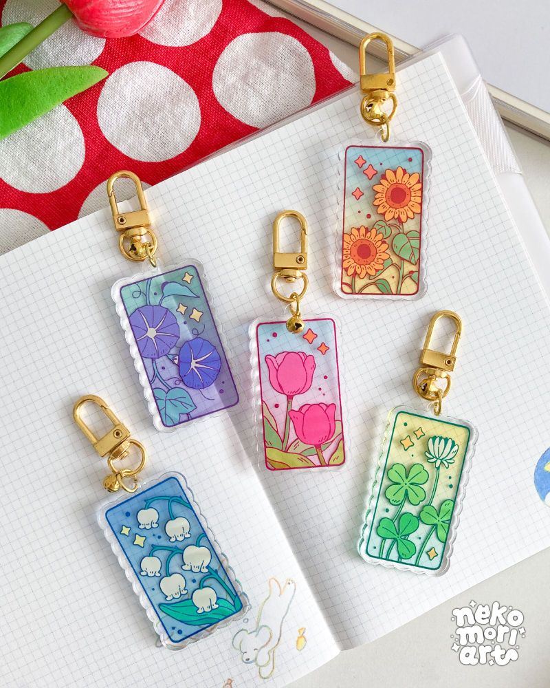 Styled product photo showing five acrylic charms on top of an open notebook. Each charm depicts a plant/flower against a translucent color background and is finished with a gold clasp and a small bell. From L-R: lilies-of-the-valley, morning glories, pink tulips, sunflowers, and clovers.