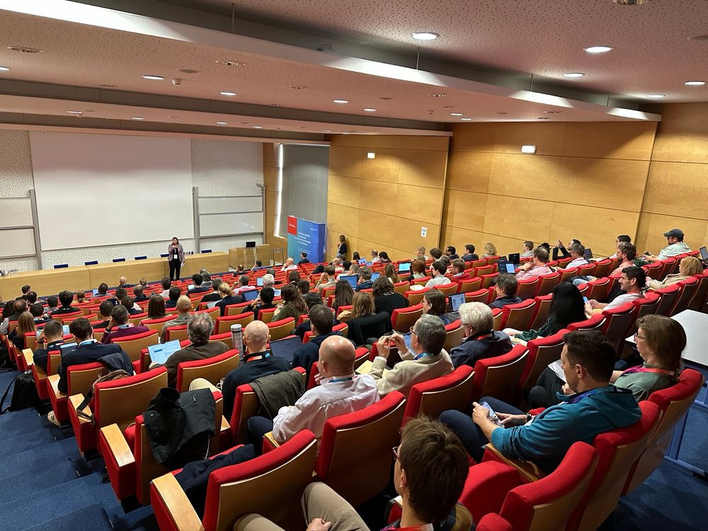 A lecture hall full of people, the picture is taken from above. In the foreground, you can see a person speaking.