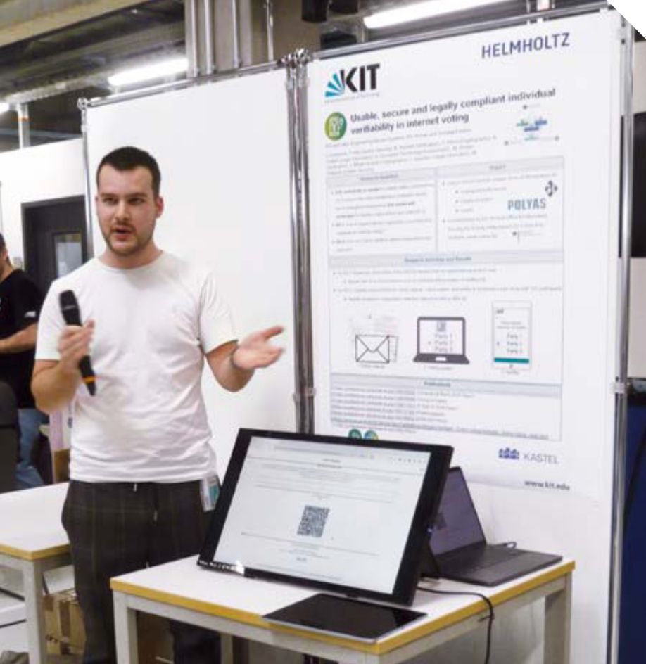 Ein Mensch steht mit einem Mikrofon in der Hand vor einem Poster und erklärt etwas. Der Inhalt des Plakats ist nicht zu lesen, die Überschrift lautet: Usable, Secure, and Legally Compliant Individual Verifiability in Internet Voting. Vor dem Poster steht ein Tisch mit einem Monitor, auf dem eine Website zu sehen ist.