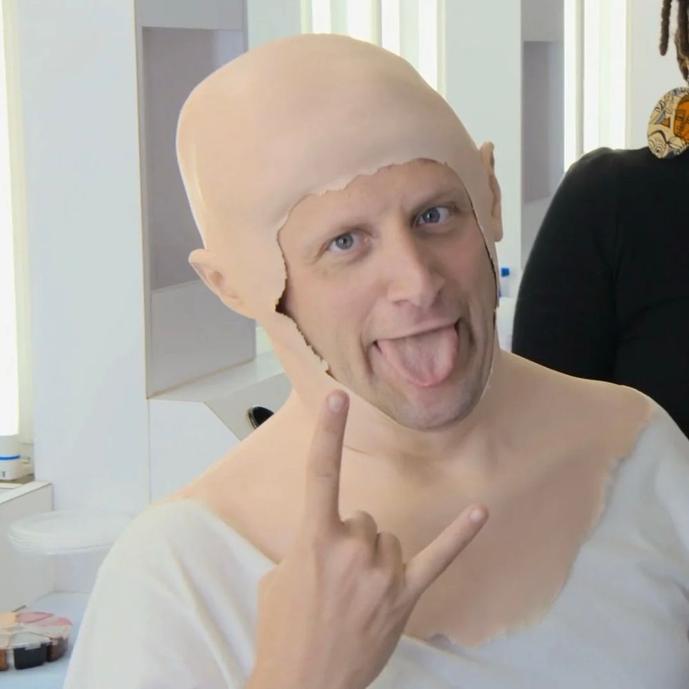 Carmine Leguzio sticking his tongue out, holding up his pinky and index finger and looking excited as he puts on prosthetic makeup to take up the mantle of Karl Havoc (from I Think You Should Leave)