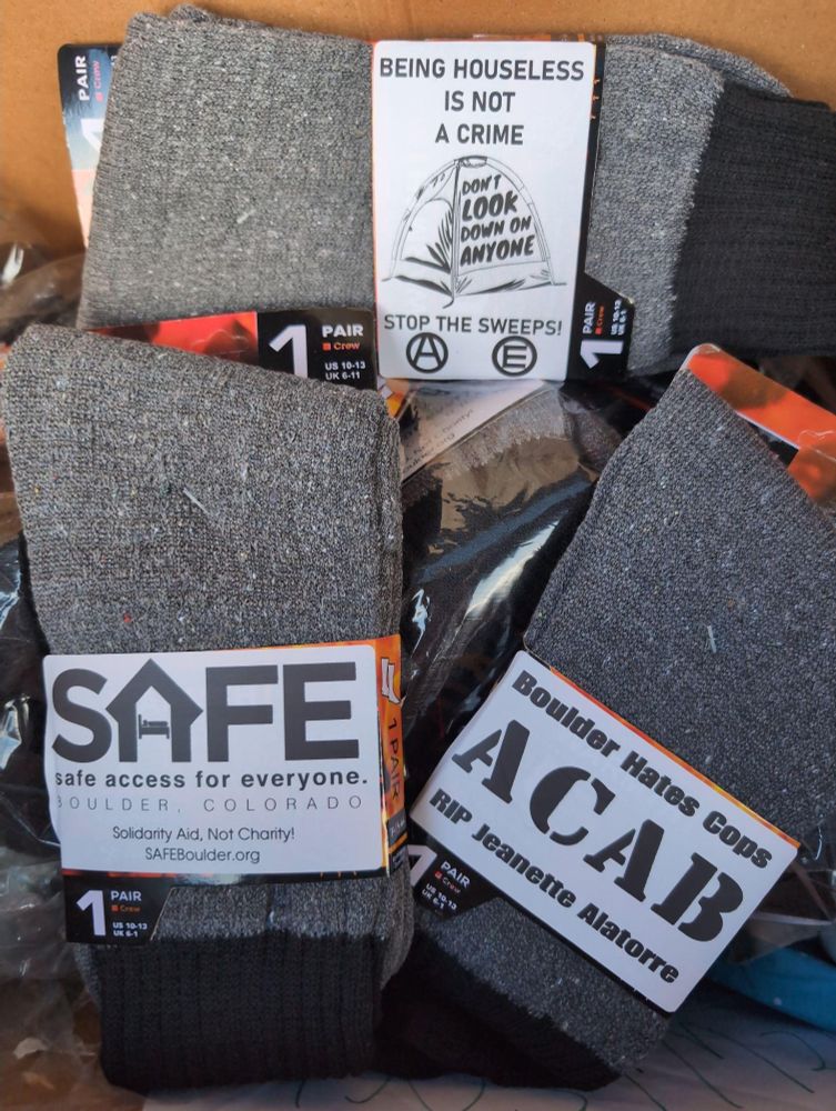 Three pairs of brand new socks. One has a SAFE Boulder sticker on it. Another says "Being Houseless is not a crime. Don't look down on anyone. Stop the sweeps" with a drawing of a tent. The last sticker says "ACAB. Boulder hates cops. RIP Jeanette Alatorre.