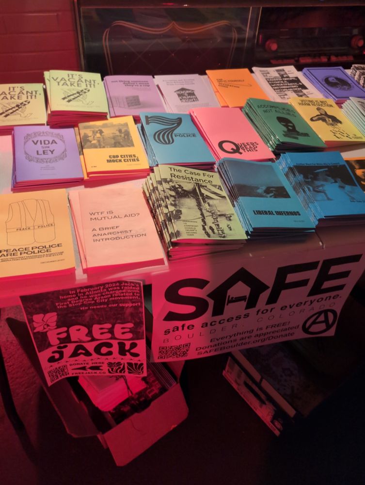 A table covered in zines. A sign with the SAFE logo is hanging off the front, and a poster to free Jack from the Stop Cop Campaign as well