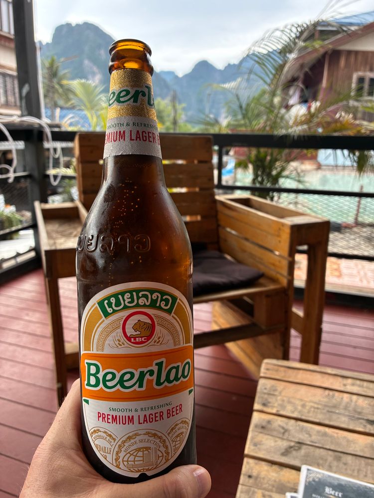 Im Bildzentrum eine 640ml Flasche Beerlao Premium Lager. Dahinter ein Palettenstuhl in einer Cocktailbar. Im Hintergrund ein bewaldeter Gebirgszug in VangVieng.
