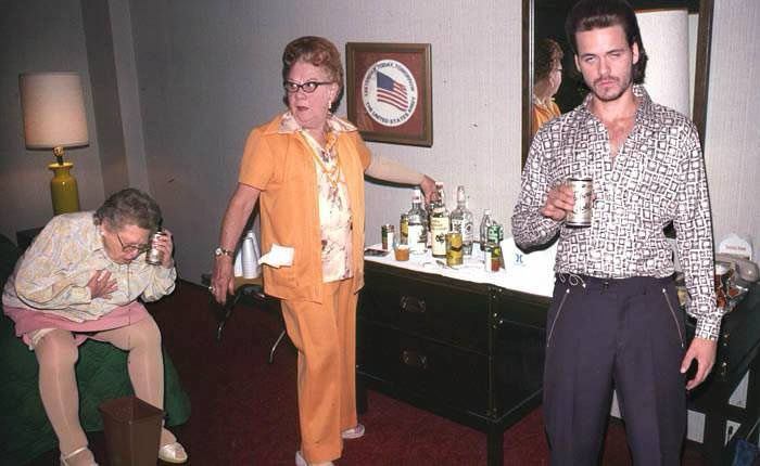 2 older women and one young man, all in front of a table of hard liquor. One older woman is sitting on the edge of the bed throwing up into a trash can