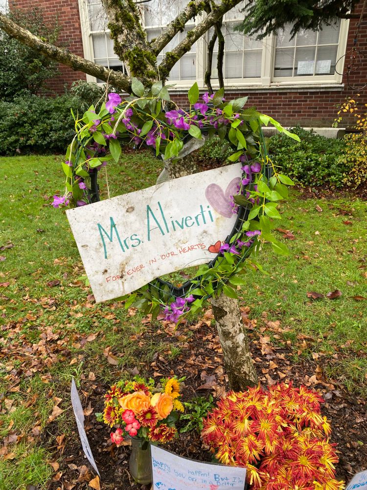 A small tree with potted flowers at its base and a floral heart hanging from a branch with a sign reading “Mrs Aliverti, always in our hearts.”