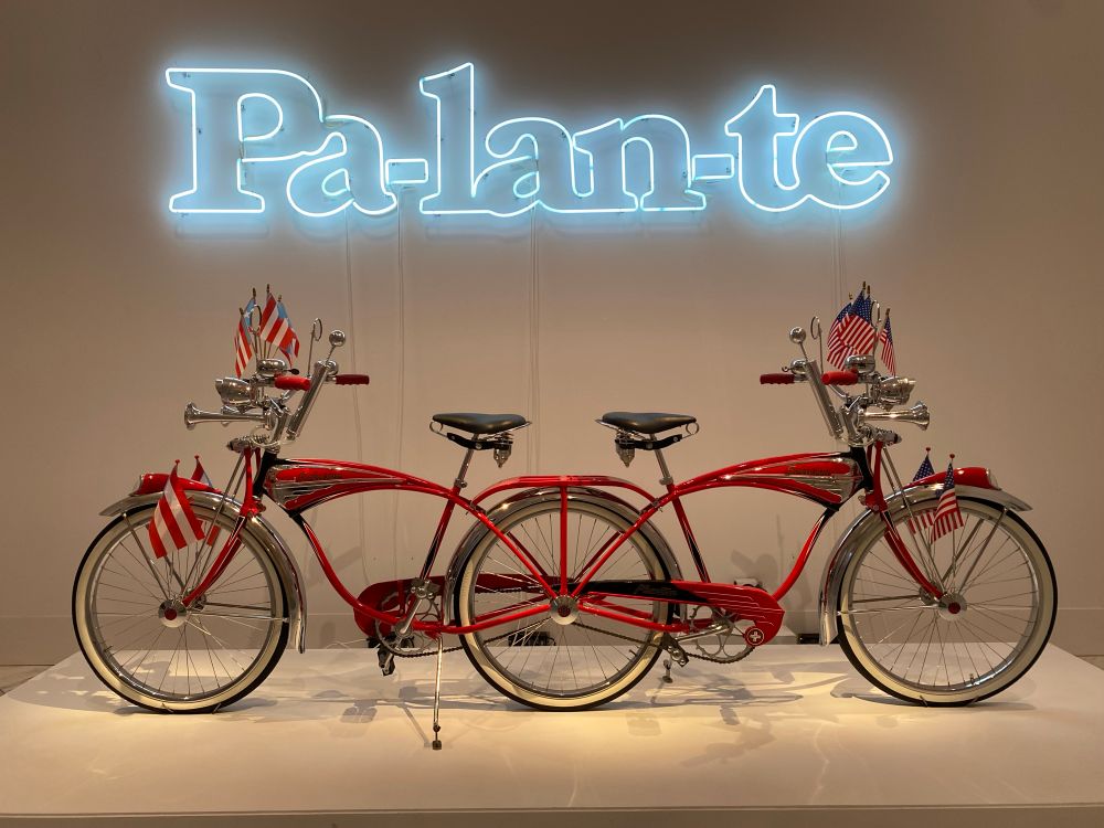 Miguel Luciano, Pa-lan-te, 2017. Two cherry-red Schwinn bicycles, decked out with horns and American and Puerto Rican flags, are joined to form one three-wheeled, double-headed creature. The word pa'lante, Spanish slang meaning "forward," glows above. Paired with a bicycle that can go in two directions at once--and so goes nowhere--the word also takes on an ironic meaning.