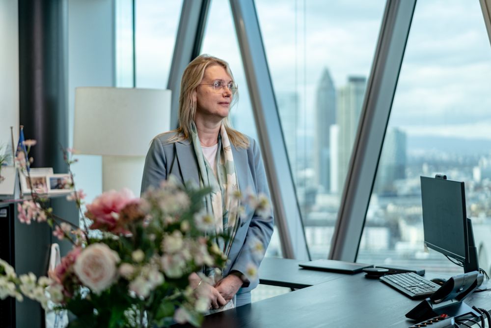 Petra Heikema, chairperson of EIOPA, stands at her desk in her office. She is wearing a light blue suit. In the background the skyline of Frankfurt am Main with the Messeturm can be seen.
