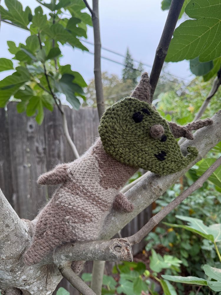 A brown and tan treelike crocheted creature with a leaf on the face with nose poking out and markings for eyes. The plushy is sitting in a fig tree tilted.