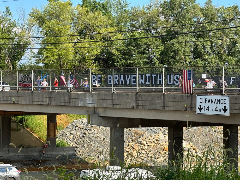 Large sign on an overpass fence reading “Be Brave with Us” and a half dozen people holding other small signs and some American flags. 