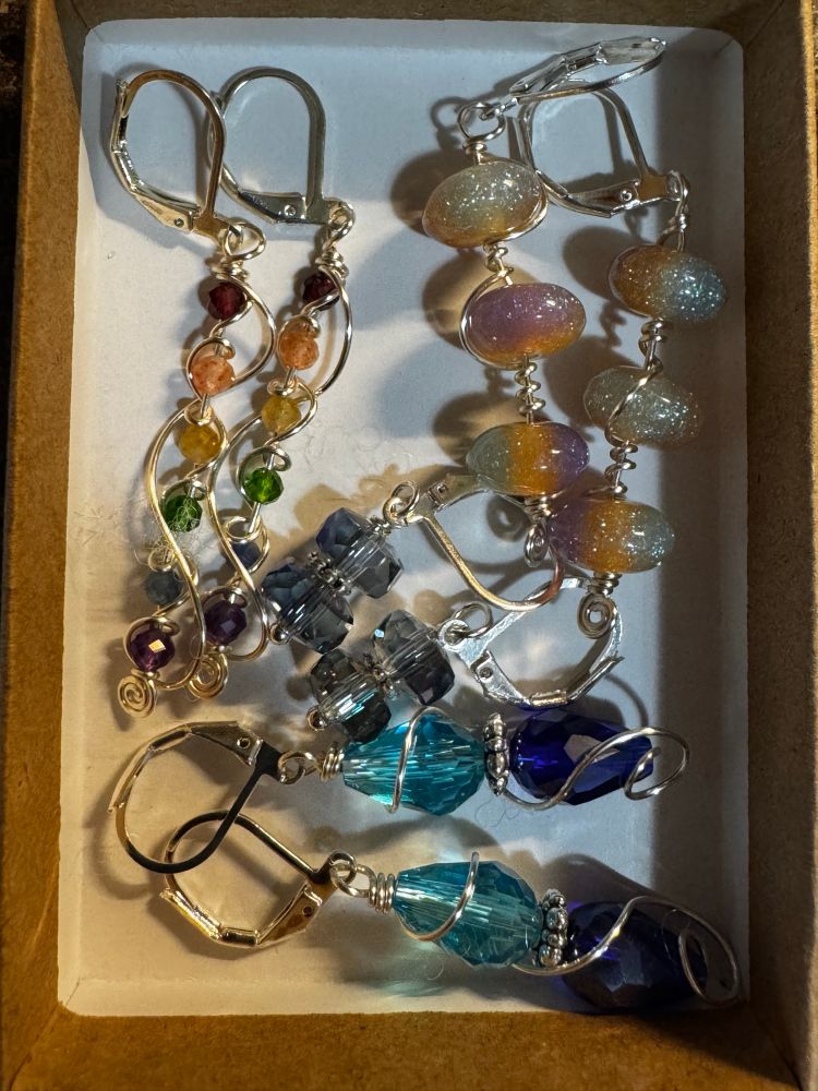 A small box of earrings: rainbow dangles, opalescent round beads in a row, blue and green beads, and small dark purple heads, all wrapped with silver wire accents. 
