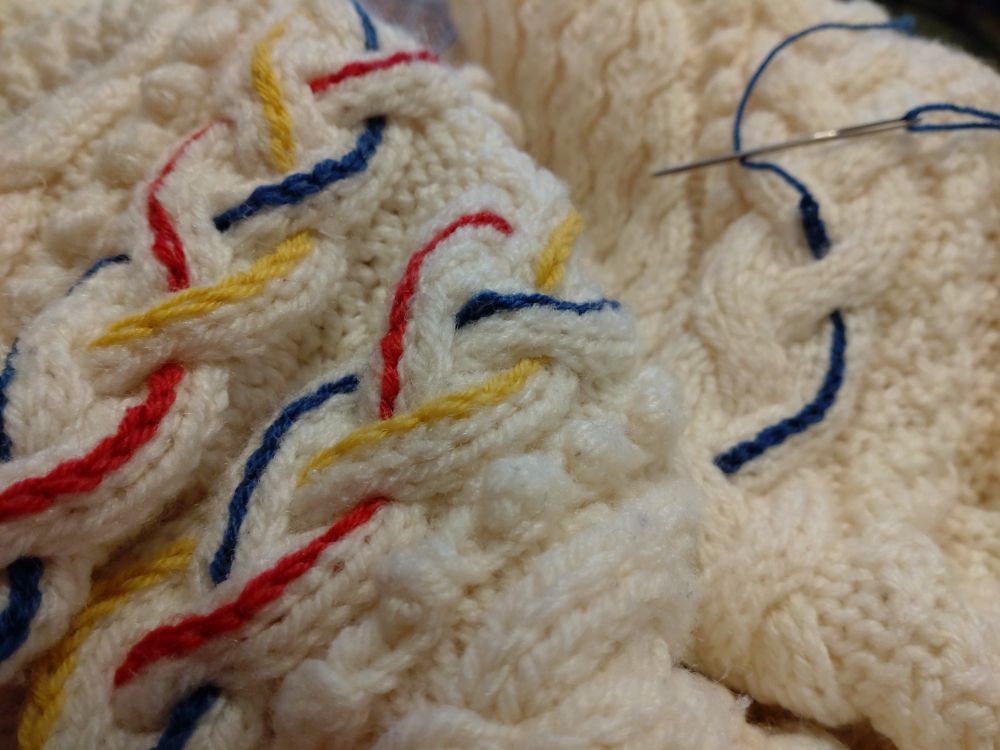 A cream Aran cardigan with embroidered chain stitching in yellow, red and blue following the cables