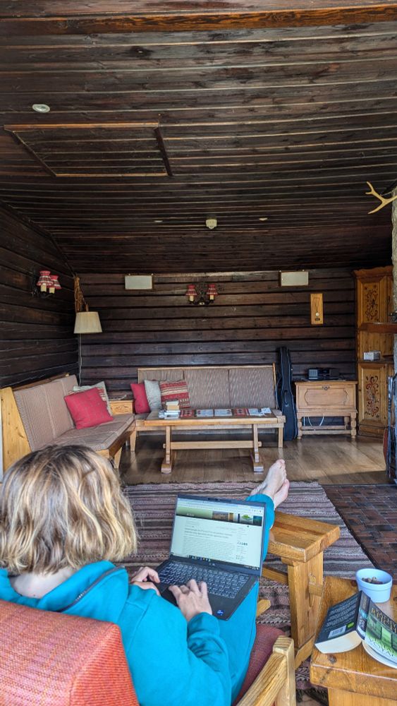 Photograph of a blonde woman wearing an aqua onesie, sitting in a red chair facing away from the camera, with her feet on a wooden footstool and a laptop open on her lap. Background is the interior of a Norwegian-style alpine hut.