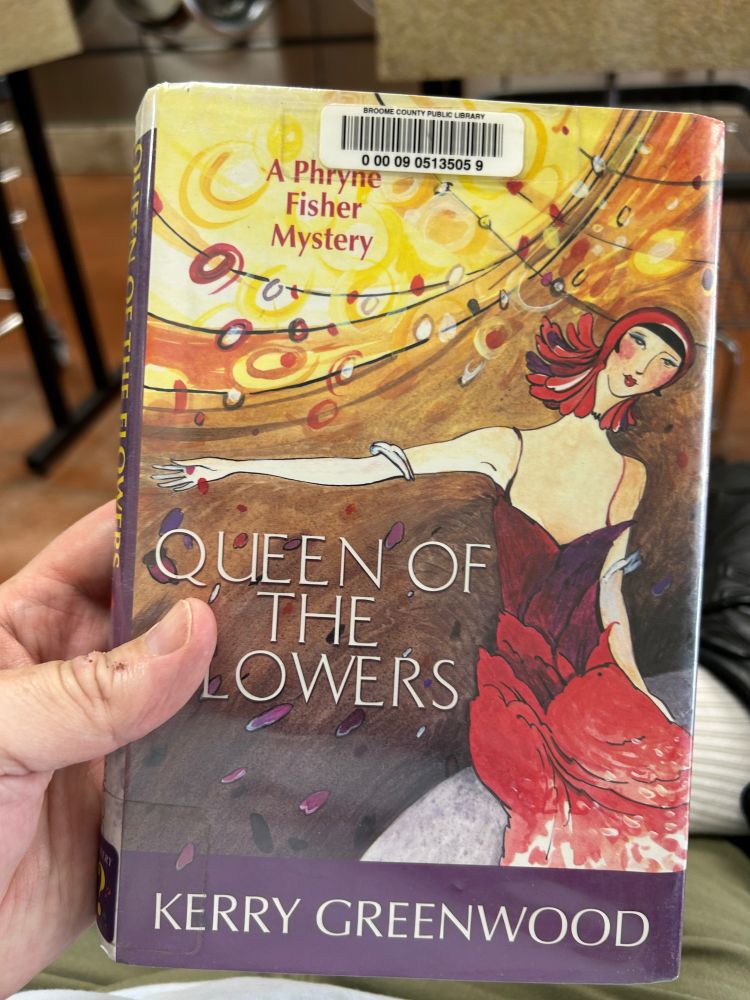 Me holding a copy of Queen of the Flowers: A Phryne Fisher Mystery at the laundromat.