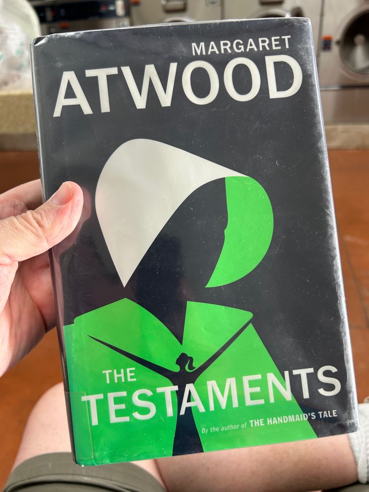 My hand holding a copy of The Testaments by Margaret Atwood.
