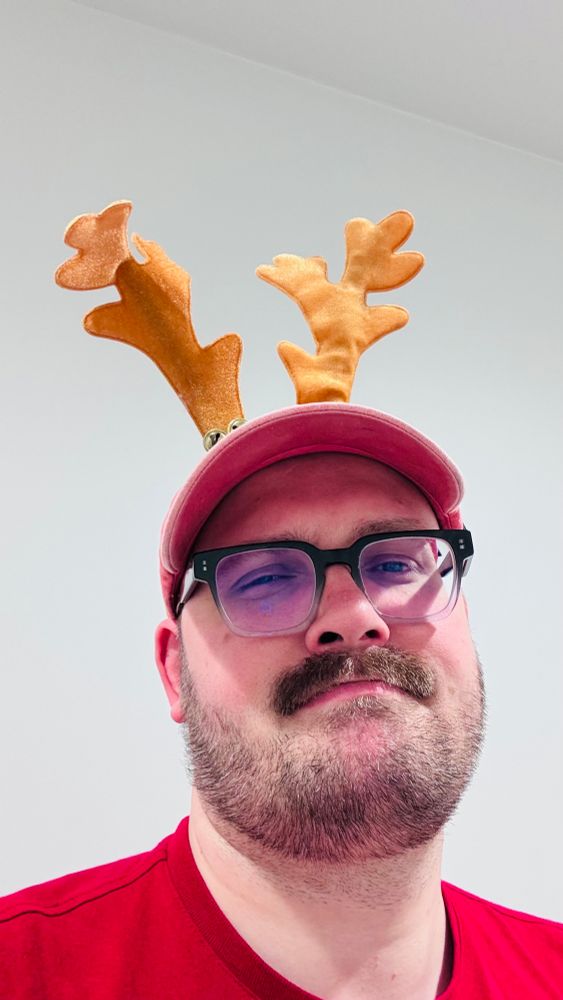 Milkydiscodick looking directly into the camera, slightly winking his right eye, from a lower angle. He is wearing a pair of fuzzy Christmas reindeer antlers on top of his head. He has a lovely short moustache, is wearing a pink sports cap & a red T-shirt. 