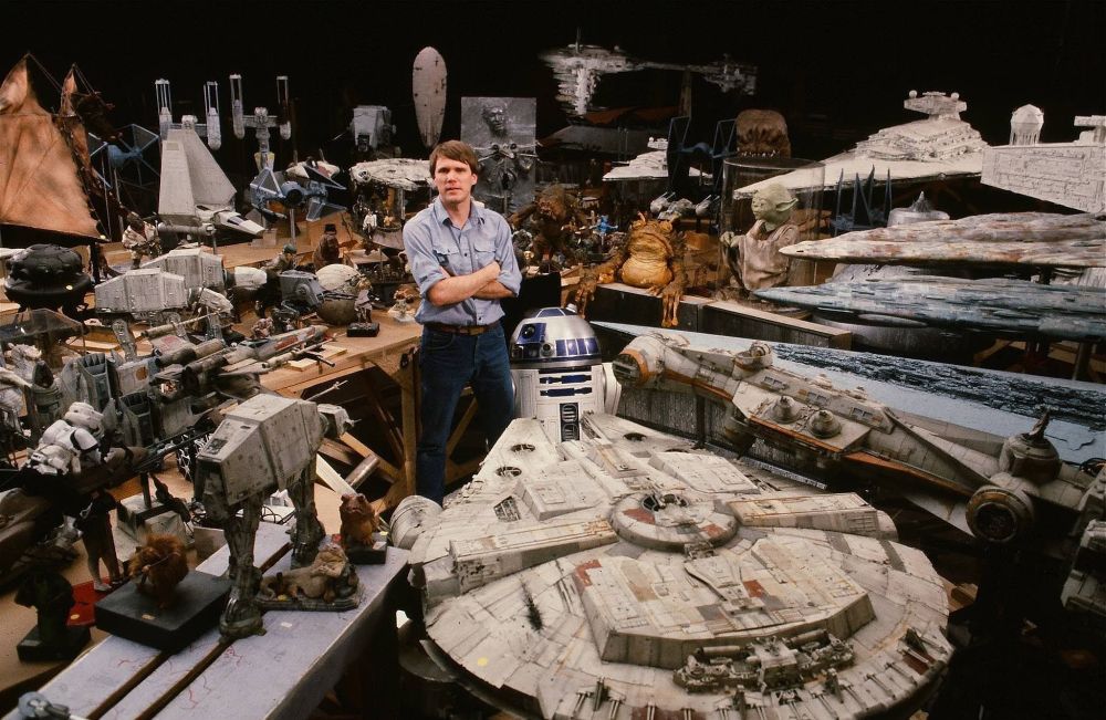 A wide shot shows Joe Johnston, a visual effects artist, standing with his arms crossed in a workshop filled with numerous models and props from the Star Wars films. He is a middle-aged man with short brown hair, wearing a light blue button-down shirt and blue jeans. He stands slightly behind a large model of the Millennium Falcon, which is prominently displayed in the foreground (upside down, probably to conceal its support).

Surrounding him and the Millennium Falcon are various other iconic vehicles and characters from the Star Wars universe, including models of X-wing fighters, TIE fighters, AT-AT walkers, and figures of Yoda and Jabba the Hutt. The models are intricately detailed and show signs of weathering and use, suggesting they were used in the filming of the movies. The workshop appears cluttered but organized, with models placed on tables and shelves, illuminated by studio lighting. The background is dark, drawing focus to the collection of Star Wars memorabilia and the artist.