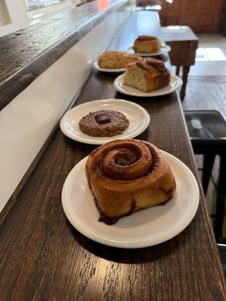 An array of five pastries on plates on a skinny wooden bar: a cinnamon roll, a chocolate chunk cookie, a cheesy pesto roll, a savory scone, and another cinnamon roll