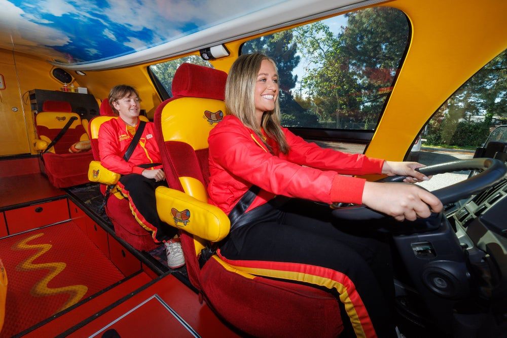 A woman sitting behind the wheel of the Oscar Mayer Weinermobile, a groovy, 70s-like interior that's mostly ketchup red with splashes and swirls of mustard yellow and a roof painted like it's a cloudy sky