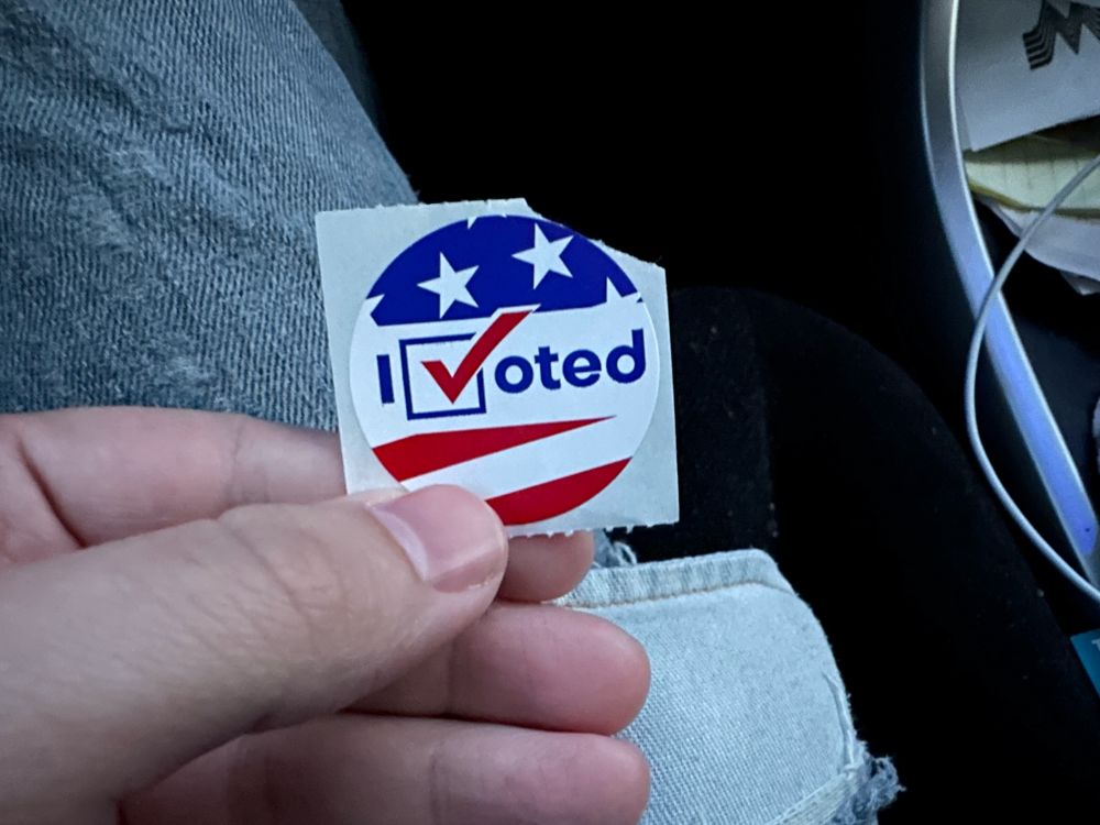 A close up image of an “I Voted” sticker, held between a thumb and forefinger.