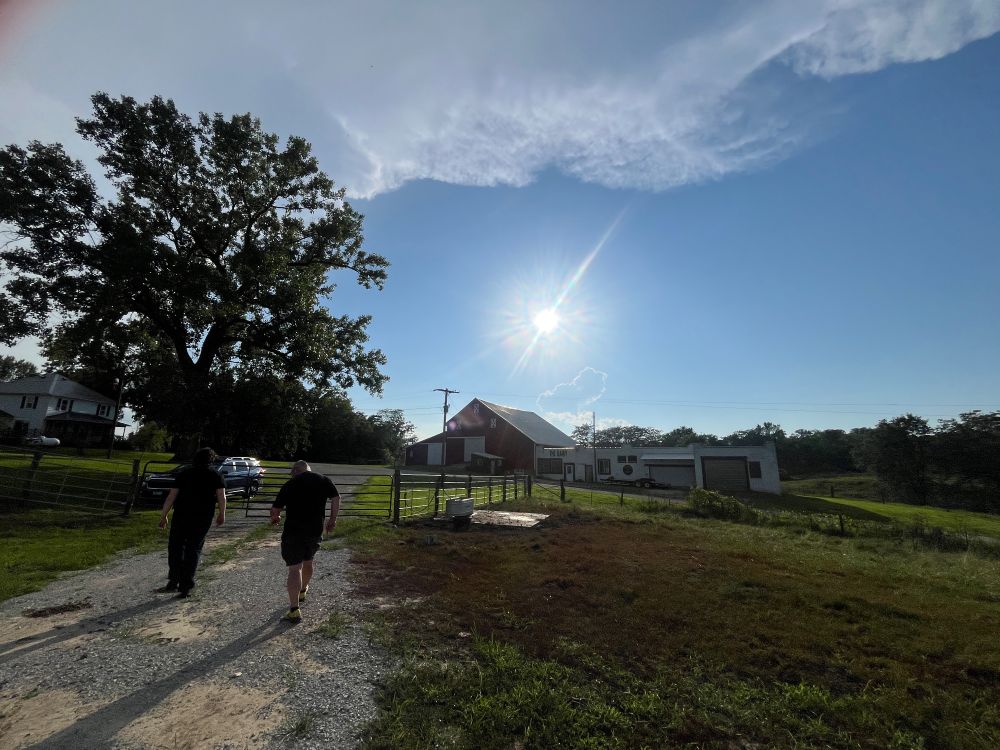 Walking back to the car in the steamy heat. A gravel path on the left with two people walking away, a fence in front of them, large tree with clouds overhead, sun blazing in the center, a barn and farm buildings in the distance. 