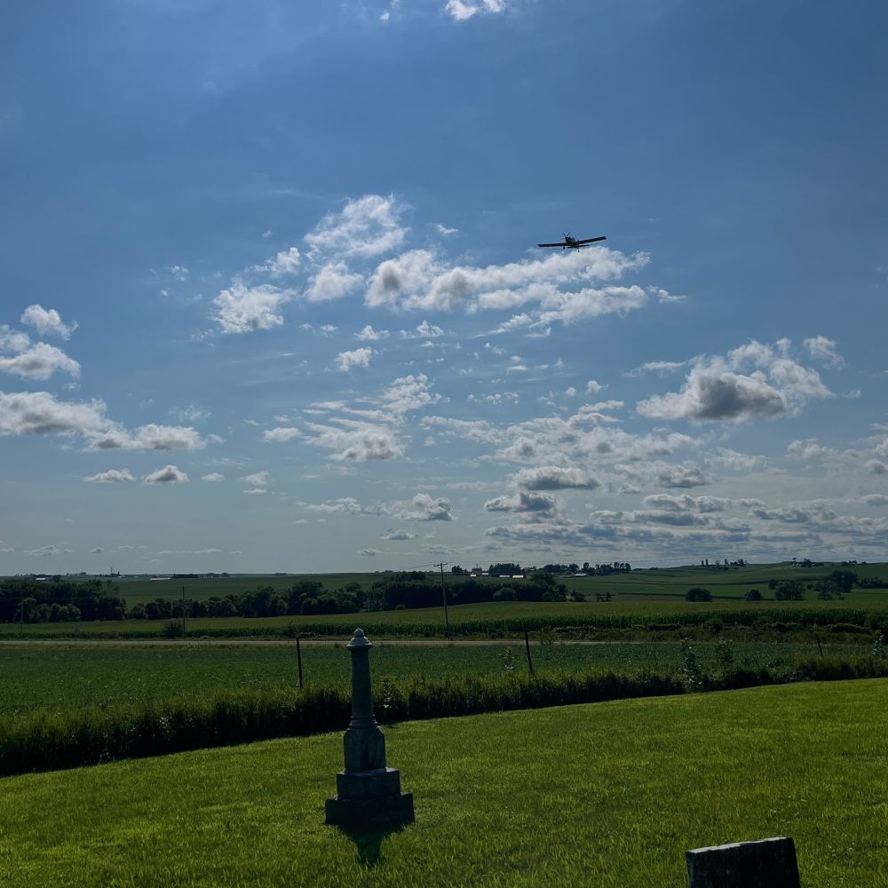 A cemetery in the foreground, a small airplane in the party cloudy sky. 