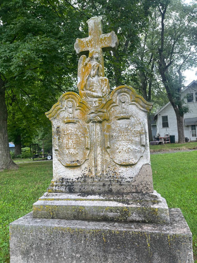 This was a different cemetery in Illinois, Edwardsville. A large stone with a cross at the top, a praying angel figure, resting atop a double headstone for Stokes and Reynolds. With hands clasped symbol between the stones. Most of the text is illegible, but death dates are 1873 and 1875, respectively. Large green trees and a White House in the background. 