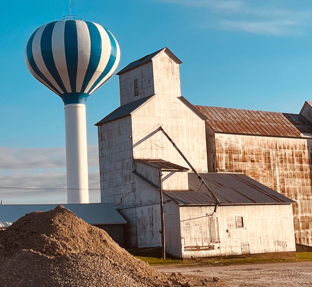 A photo of a grain elevator and out buildings in shades of white and rust. To the left is a water tower, taller than the other buildings, striped blue and white, looking very much like a hot air balloon. In the foreground to the left is a big pile of sand. Light blue sky in the background with a few low clouds. 