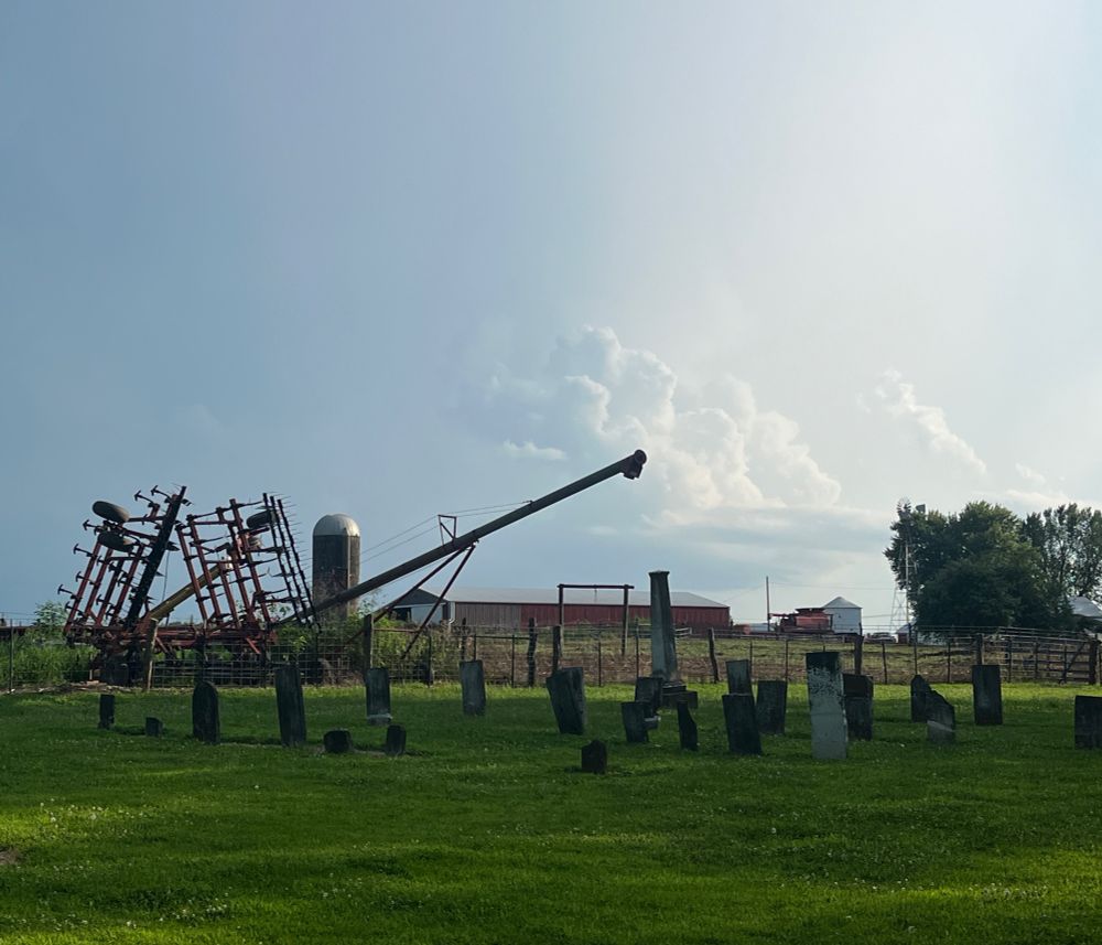 Southeast Iowa pioneer cemetery - in the foreground a field with headstones in rows, farm implements beyond, including a grain auger looming overhead, with storm clouds in the background highlighted by the sun. 
We pulled up and the farmer drove across the grass to block our car. These places are public and protected by law. He didn’t make it easy to get in, but we did (my partner has great people skills). After 2 gates, with 4 chains/ties, we walked in. This cemetery was called “Cholera Hill” because of an outbreak in 1851. 