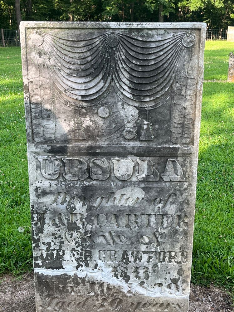 A large weathered rectangular headstone reads, “URSULA daughter of J&B CARTER wife of JOHNE CRAWFORD Nov 16, 1849 aged 30 years” Above the text is carved imagery, parted curtains, flowers, an urn. 