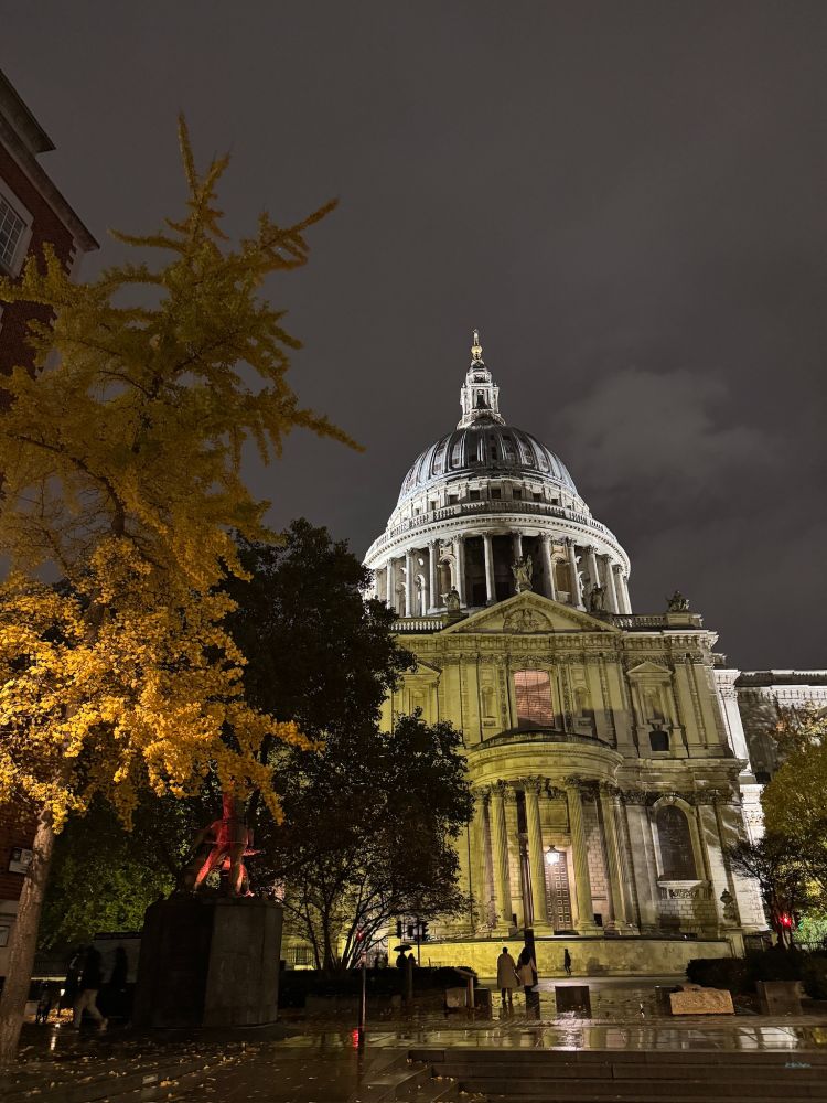 A quiet, rainy night outside St Paul’s Cathedral in London. The dome glows softly against the dark sky, autumn leaves shimmering in the streetlights while a few late walkers cross the wet square.
