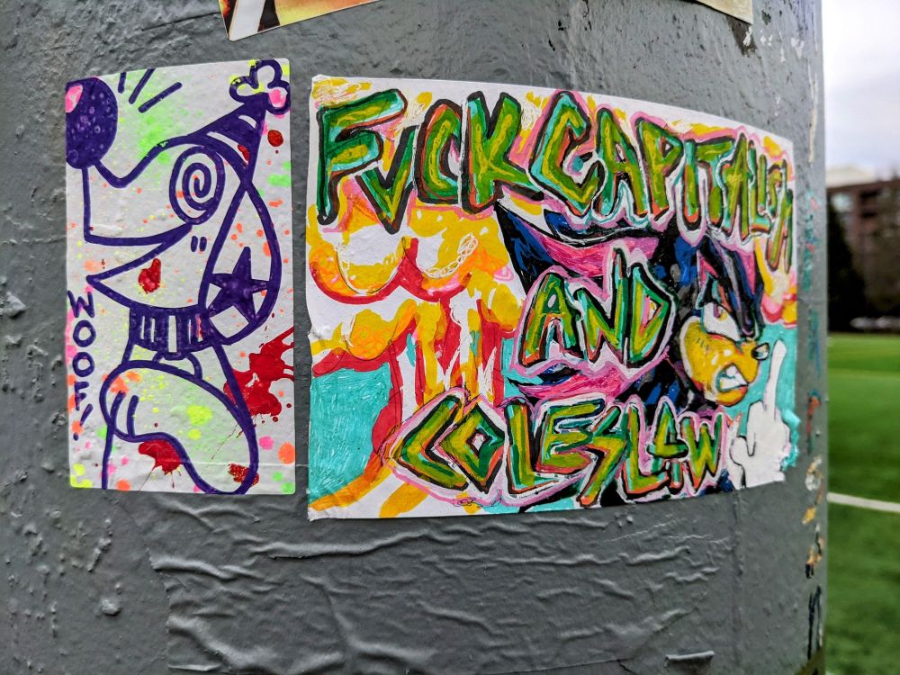 Photo from my collection, dated early 2023 in cal anderson park. Two stickers by two different artists on a pole. A small clown dog splattered in various colors. A big awesome super cool sticker of shadow the hedgehog that says Fuck Capitalism and Coleslaw with shadow flipping the bird and a huge explosion in the background