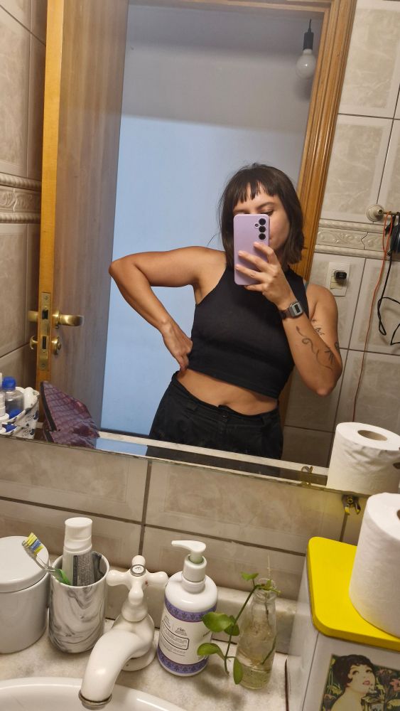 Foto minha no espelho do banheiro, mulher branca de cabelo curto e preto e franja com um top preto e calça preta fazendo pose com um braço pra baixo e outro segurando o celular