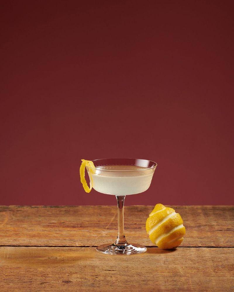 A cocktail in a coupe glass garnished with a lemon twist, set on a wooden table with a whole lemon beside it against a maroon background.
