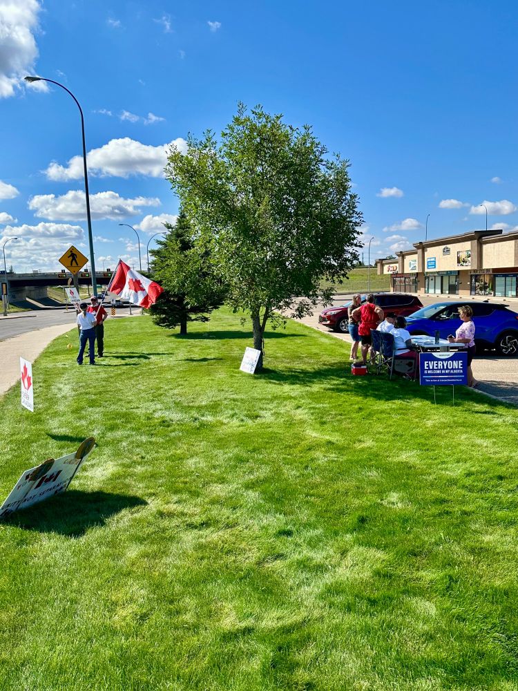 Photo of the people gathering signatures at the Justin Wrights UCP office venue. Two people are standing on the edge a grass area beside thirteenth avenue, my wife holding a Canadian Flag and @katfromthehat.bsky.social holding a ForeverCanadian sign attached to a hockey stick. There are also lawn signs informing people they can sign the petition at this location. on the parking lot side of the grass area is a table with two people taking signatures. Other people are there to sign the petition, or discussing the petition. 
