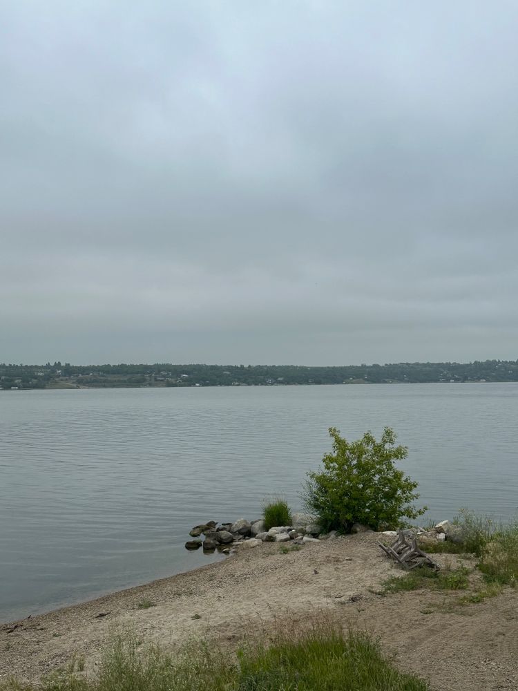 Photo from the shoreline along Buena Vista Beach, one of the four major settlements along Last Mountain Lake. The others being Regina Beach, Saskatchewan Beach and Wee Too Beach. In the foreground is some grass along the edge of a sandy beach; a bench that was turned over during a recent wind storm; a makeshift rock pier and a green bush growing at the edge of the beach. In the background on the far side of the lake there are cabins and homes that have been built along the edge of the lake.  The sky is cloudy and overcast. Much of our time was spent indoors. 