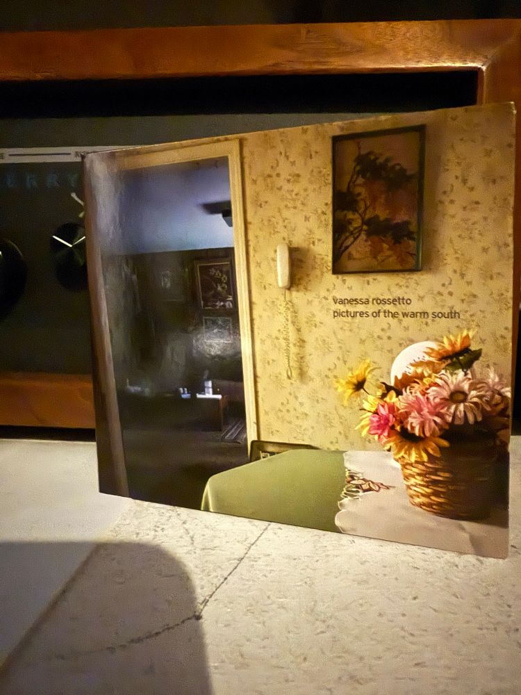 CD sleeve art showing a household scene with a basket of zinnia flowers on a table; yellow-and-green floral-print wallpaper; a light-yellow landline phone mounted on the wall, and a view through a doorway into a darker room. The CD sleeve leans against a stereo component with a matte-black face and shiny black dials.
