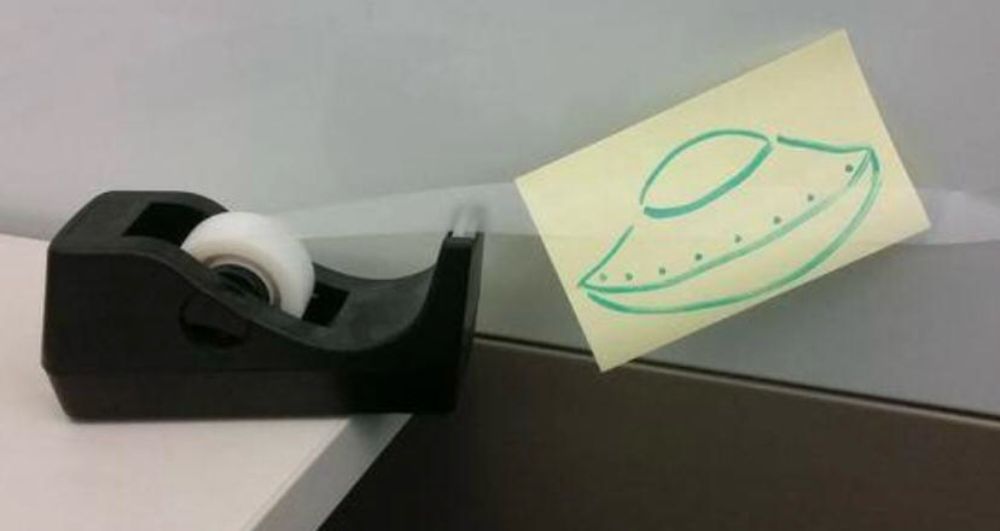 “UFO caught on tape” - Image of a post it note with a simple alien spaceship drawing attached to a dry erase board with a piece of tape from a desktop tape dispenser. 