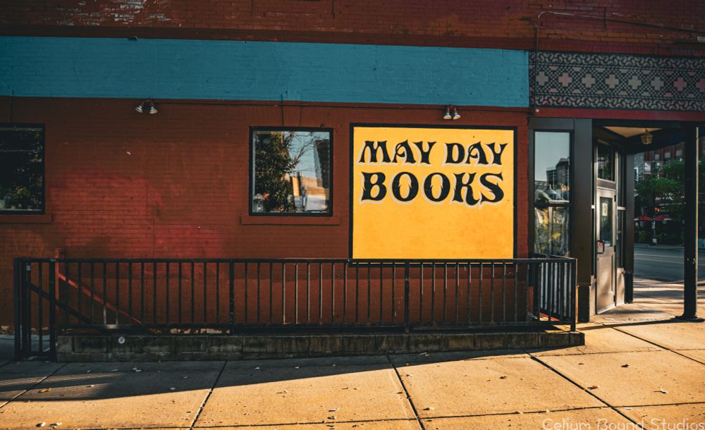 The side view of the store, which features a red brick building with a large blue strip above two windows, and a yellow square sign with black writing  that says,"May Day Books." A stairwell that leads below the street level and its fence can be seen below the sign and along the wall. 
