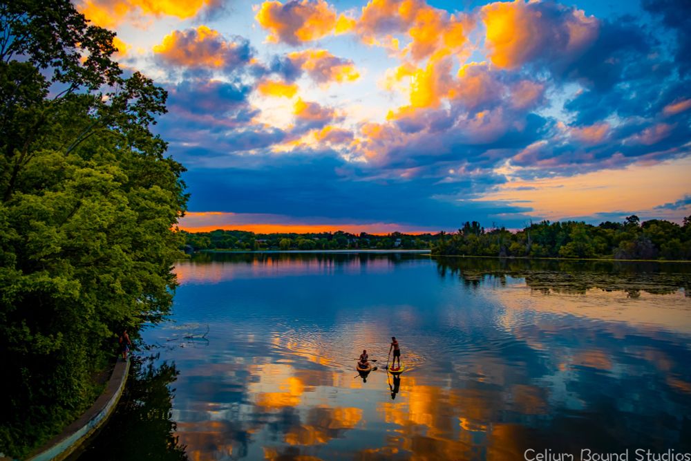 A vibrant sunset over Lake of the Isles in Minneapolis. The sky is shades of orange, yellow, pink and blue and all of it is reflected off the water. In the middle of the reflection two paddles boarders, one sitting and one standing, can be seen. The trees of Minneapolis line the shores in every direction. 
