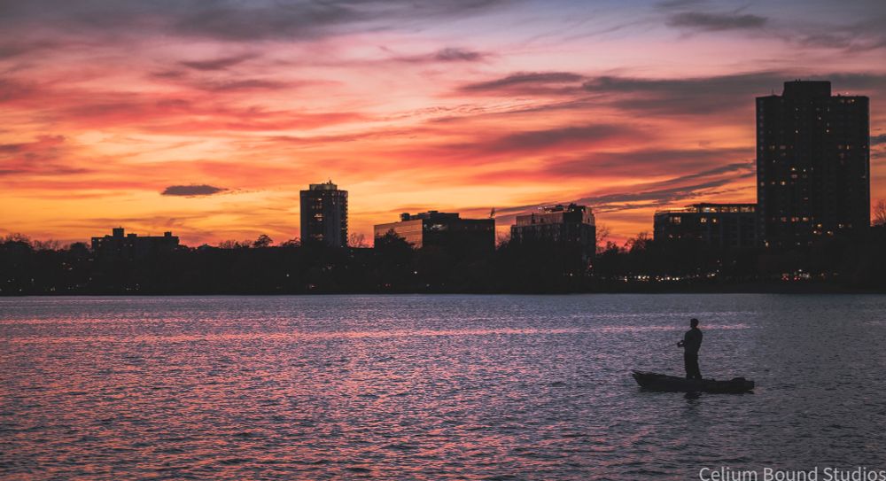 A man stands on a fishing kayak in a lake, sunset in the distance with a skyline of skyscrapers and trees. 