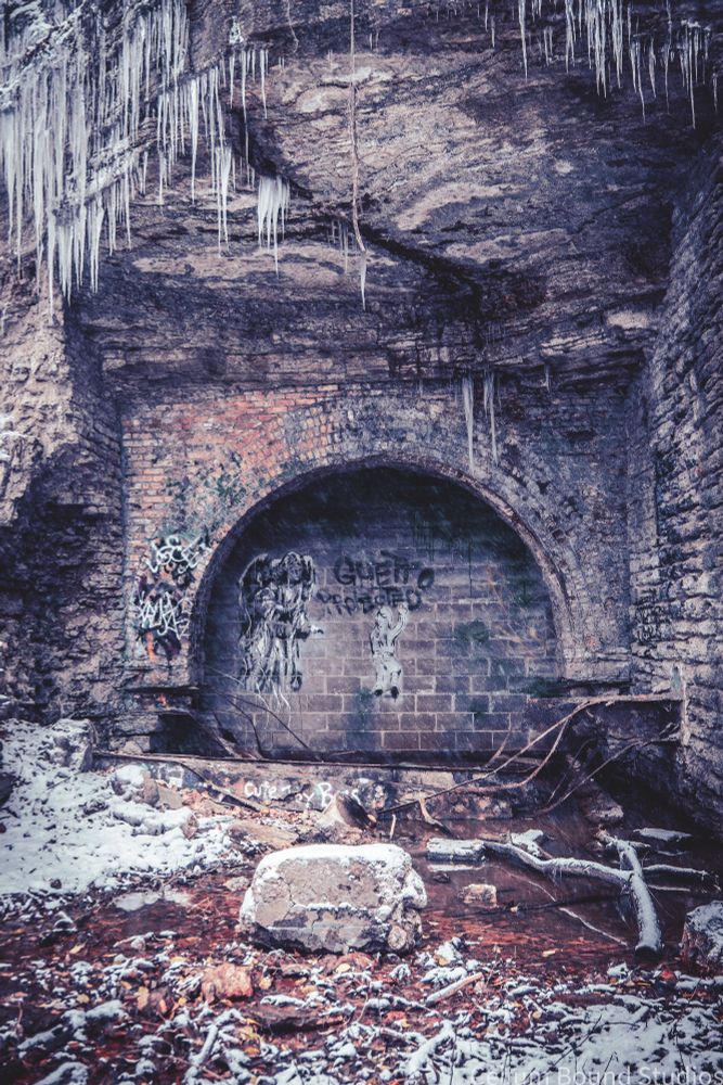The ruins of area of an old mill, graffiti covers the brick. The ruin is recessed in the side of a stone hill, icicles hang above