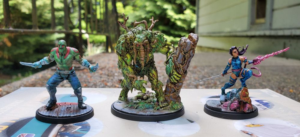 Painted miniatures for the game Marvel Crisis Protocol. Drax, Man-Thing, and Psylocke. The picture is taken outdoors with a house and trees in the background.