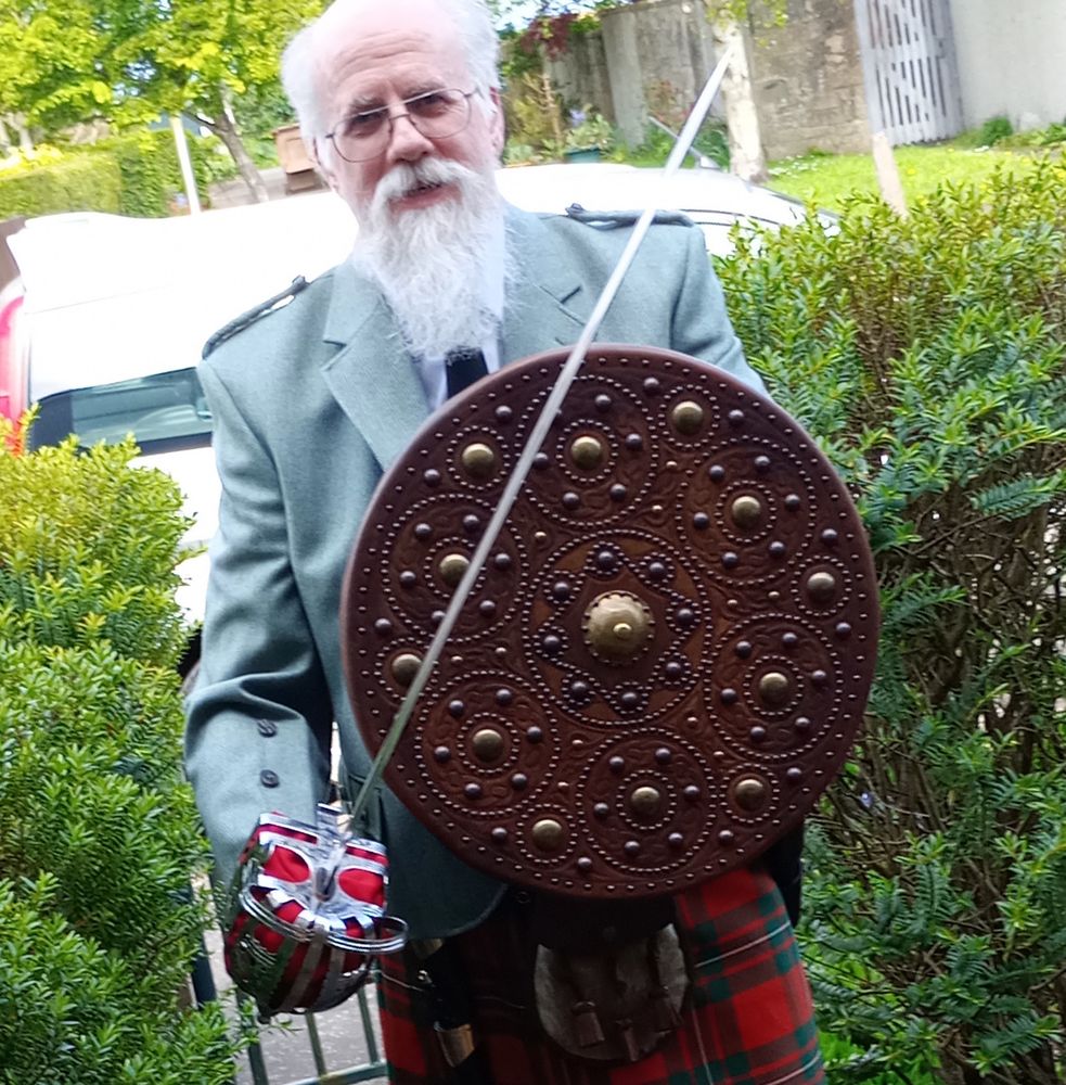 A man with a white beard is wearing a modern kilt and jacket, while carrying a basket-hilted broadsword and a targe. The hilt of a dirk is visible at his waist on his right, while the top of the sword’s scabbard is just visible on his left hip. It’s on a suburban garden path with bushes on either side and a car in the background.