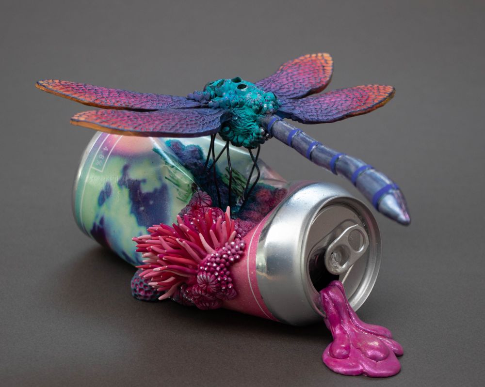 A dragonfly sculpture is sitting on a drinking can. The dragonfly is colored in a pastel rainbow ranging from a light coral, to pink, to purple to turquoise.
A pink glittery goo is running out of the drinking can.