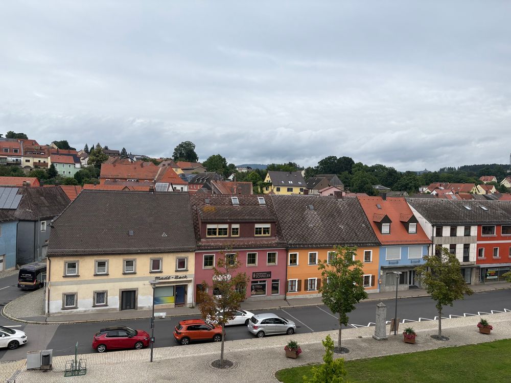 Blick über den Marktplatz und die Dächer und den Marktplatz einer bayerischen Kleinstadt. Im Vordergrund sind bunte Häuser in beige, rotbraun, orange und gelb. Im Hintergrund verliert sich der Ort hinter einer Reihe Dächern. Am Ortsrand ist bereits der Wald zu sehen.